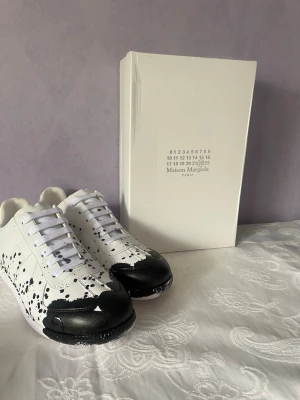 Maison Margiela vita sneakers med svart färgstänk - Maison Margiela sneakers i vitt läder med unika svarta färgstänk och svart detalj på tån. Klassisk låg modell med vita skosnören och gummisula. Snygg och konstnärlig design som sticker ut i mängden.