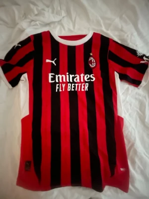 AC Milan matchtröja Puma röd/svart - Säljer en AC Milan matchtröja från Puma i rött och svart med vita detaljer. Tröjan har klassiska vertikala ränder, klubbmärke och Puma-logga på bröstet samt Emirates Fly Better-tryck. Tillverkad i lätt och ventilerande polyester, perfekt för fotboll.