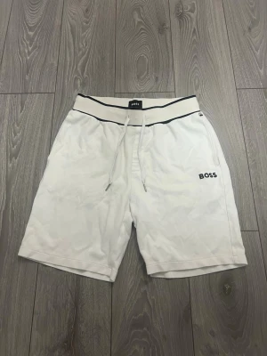 Vita shorts med snörning från BOSS - Snygga vita shorts från BOSS med svart logga på benet och snörning i midjan. Shortsen har en relaxed passform och elastisk midja med kontrastfärgade detaljer. Perfekta för sommardagar och chill häng.