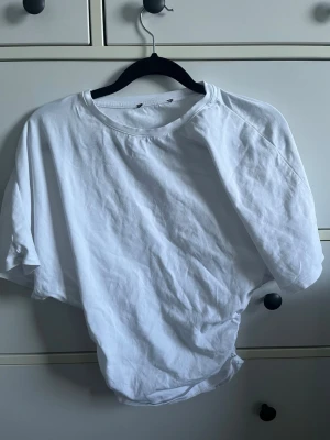 Vit t-shirt med snörning i sidan - Säljer en vit t-shirt i bomull med korta ärmar och rund hals. T-shirten har en form av scrunch på sidan vilket gör den lite tightare längst ner i passformen. Perfekt till jeans eller kjol för en avslappnad stil.
