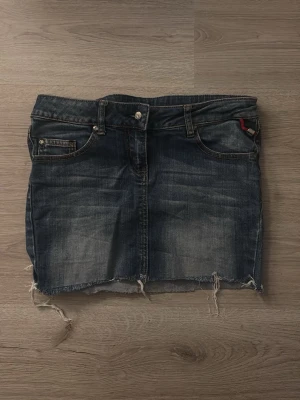 Mörk jeanskjol från Tally Weijl - Snygg mörkblå jeanskjol från Tally Weijl med slitna kanter och dekorativa paljettdetaljer på bakfickorna. Klassisk femficksmodell med knapp och dragkedja framtill. Perfekt för dig som gillar en edgy och trendig stil.