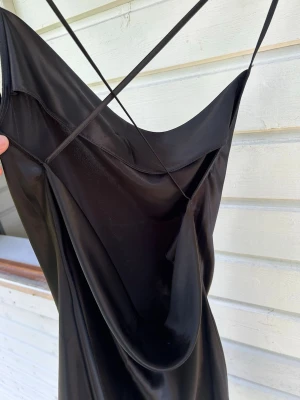 Svart långklänning med tunna band - Säljer en elegant svart långklänning med tunna axelband som korsas i ryggen. Klänningen har en draperad urringning och är tillverkad i ett glansigt material som ger en lyxig känsla. Perfekt för dig som vill ha en stilren och trendig look. 