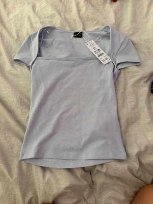Ljusblå kortärmad topp - Säljer en ljusblå topp med korta ärmar och rak halsringning. Toppen har en enkel och stilren design, perfekt att matcha med jeans eller kjol. Tillverkad i mjukt bomullsmaterial för en bekväm känsla.