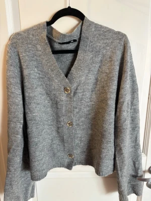 Grå stickad kofta med knappar - Mysig grå stickad kofta med tre stora knappar framtill och v-ringad hals. Koftan har lång ärm och rak passform, perfekt att slänga på sig över en t-shirt. Materialet är  mjukt och lite luddigt.
