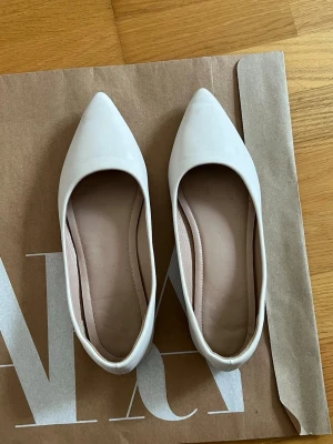 Vita ballerinaskor med spetsig tå - Snygga vita ballerinaskor med spetsig tå och låg klack. Skorna har en stilren design och är tillverkade i ett slätt material som ger en elegant look. Perfekta för dig som gillar minimalistisk stil och vill ha ett par klassiska skor till garderoben.