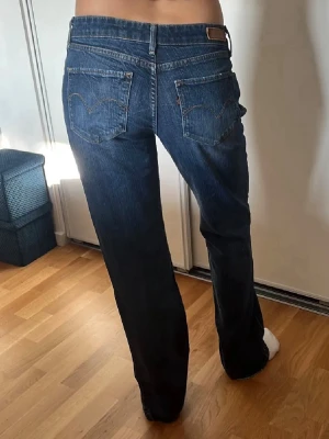 Blå Levis jeans med slitningar - Säljer ett par blå  jeans med låg midja och snygga slitningar framtill. Jeansen har klassiska fem fickor och kontrastsömmar. Mörkblå Levís jeans! Lågmidjade! Innerbensmått - 82cm Midjemåttet- 83 cm Längd - 105