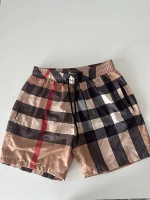 Rutiga Bur berry shorts med snörning - Snygga shorts med klassiskt rutigt mönster i beige, svart, vitt och rött. De har elastisk midja med svart snörning och sidofickor. Perfekta för sommardagar och ger en trendig vibe till din outfit. Små i storleken!