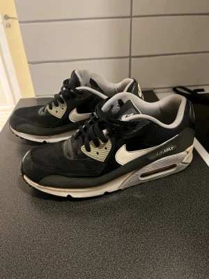 Svarta Nike Air Max sneakers - Säljer ett par svarta Nike Air Max sneakers med vita detaljer och grå insida. Skorna har en klassisk låg siluett, mesh och syntetmaterial i ovandelen samt den ikoniska Air Max-sulan för extra komfort. Perfekta för dig som gillar streetwear och sportig stil.