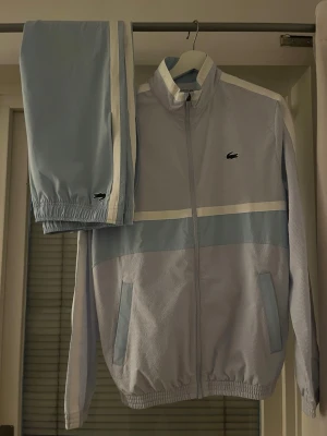 Ljusblått träningsset från Lacoste - Säljer ett stilrent ljusblått träningsset från Lacoste med vita detaljer. Jätte rent använt och passar M/L