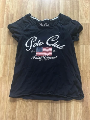 Svart Polo Club t-shirt med tryck - Svart t-shirt från Polo Club med stort vitt tryck på bröstet och amerikansk flagga. Rund halsringning och korta ärmar. Vill du ha fler bilder på plagget? Kontakta mig så fixar jag! Den har använts en gång. 