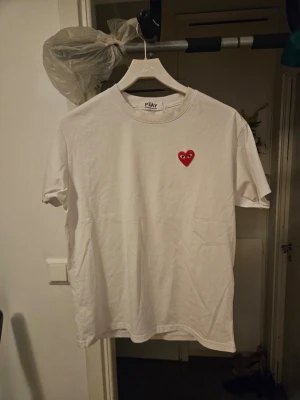 Vit t-shirt Comme des Garçons Play - Vit t-shirt från Comme des Garçons Play med det ikoniska röda hjärtat med ögon broderat på bröstet. Klassisk passform och rund halsringning. Perfekt basic-plagg med en twist som funkar till allt.