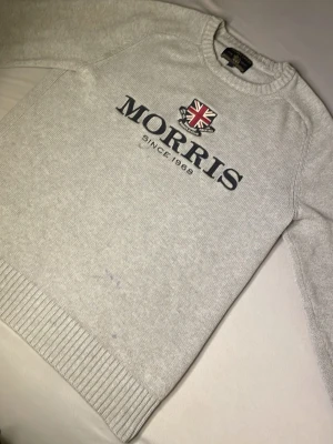 Ljusgrå tröja från Morris - Ljusgrå stickad tröja från Morris med rund halsringning och ribbade muddar. Framtill finns ett tryck med Morris-logga och brittisk flagga samt texten 'Since 1968'. Perfekt för en clean och klassisk stil.