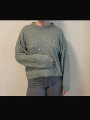 Grågrön stickad tröja med pärlor - Mysig stickad tröja i grågrönt med dekorativa pärlor över hela framsidan. Tröjan har en avslappnad passform, ribbade muddar och en högre rund halsringning. Perfekt för dig som gillar en enkel men ändå unik look.