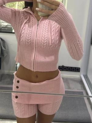 Rosa stickat loungewear-set med dragkedja - Mysigt rosa loungewear-set i stickad kvalitet med kabelstickat mönster. Setet består av en croppad hoodie med dragkedja och långa ärmar samt matchande shorts med knappdetaljer och hög midja. Perfekt för chill dagar hemma.