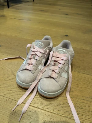 Adidas rosa sneakers med mocka - Snygga Adidas sneakers i ljusrosa mocka med klassiska vita ränder på sidorna och matchande rosa skosnören. Skorna har en låg profil och en vit sula med gummidetaljer. Perfekta för dig som gillar en clean och sportig look.