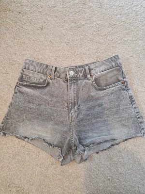 Grå jeansshorts med rå kant - Säljer ett par korta grå jeansshorts med hög midja och rå, fransig kant nedtill. Shortsen har klassisk femficksdesign, bälteshällor och knappgylf. Perfekta för en avslappnad look.