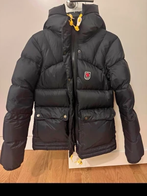Fjällräven jacka down lite women  - Snygg svart dunjacka från Fjällräven med huva och gula detaljer på snörningen. Jackan har två stora fickor med knappar framtill och en klassisk Fjällräven-logga på bröstet. Perfekt för kalla vinterdagar tack vare det tjocka dunfodret, fler bilder finns vid intresse🤎