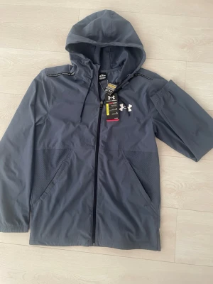 Mörkblå vindjacka från Under Armour - Snygg mörkblå vindjacka från Under Armour med huva och dragkedja framtill. Jackan har två sidofickor, justerbar huva med snören och vit logga på bröstet. Materialet är lätt och vindtätt, perfekt för blåsiga dagar.