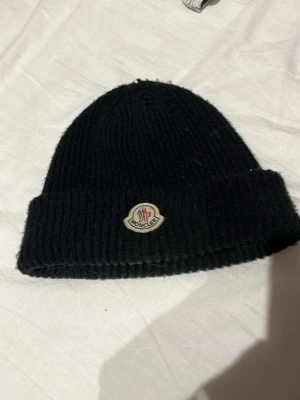 Svart ribbstickad mössa Moncler - Säljer en svart ribbstickad mössa från Moncler med uppvikt kant och klassisk Moncler-logga framtill. Mössan är i tjockt, mjukt material och har en enkel, stilren design som passar perfekt till kalla dagar.