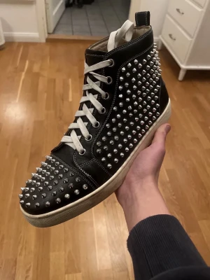 Svarta höga sneakers med nitar - Säljer ett par svarta höga sneakers från Christian Louboutin med vita snören och ikonisk röd sula. Skorna är täckta av silvriga nitar över hela ovandelen och tån, vilket ger en riktigt edgy och unik look. Tillverkade i läder med klassisk high-top design. Detta är en äkta vara.