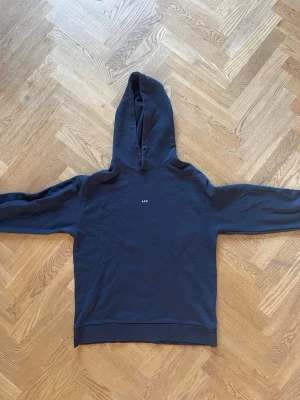 Mörkblå hoodie från A.P.C. - Säljer en stilren mörkblå hoodie från A.P.C. med liten vit logga på bröstet. Klassisk huva med snörningshål och ribbade muddar vid ärmslut och nederkant. Perfekt för en avslappnad och trendig look.