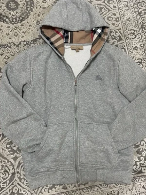 Grå hoodie med dragkedja och Burberry-mönster - Snygg grå hoodie från Burberry med dragkedja framtill och klassiskt rutigt Burberry-mönster i huvan och längs insidan. Tillverkad i mjukt material, har två fickor fram och ribbade muddar. Perfekt för en chill och stilren look.