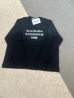 Svart långärmad topp Acne Studios - Svart långärmad topp från Acne Studios med vit text 'Acne Studios STOCKHOLM 1996' på bröstet. Klassisk rak passform och rund halsringning. Perfekt för dig som gillar stilrena och trendiga plagg.