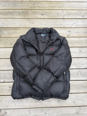 Svart pufferjacka från Polo Ralph Lauren - Säljer en svart pufferjacka från Polo Ralph Lauren med röd logga på bröstet. Jackan har hög krage, dragkedja framtill och två sidofickor med dragkedja. Tjockt vadderad för extra värme och perfekt för kalla vinterdagar.