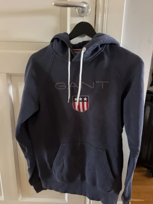Mörkblå hoodie från GANT med tryck - Mörkblå hoodie från GANT med vit dragsko i huvan och ett broderat GANT-tryck samt sköld med stjärnor och ränder på bröstet. Klassisk känguruficka framtill och mjukt material som känns skönt mot huden. Perfekt för en avslappnad och stilren look.