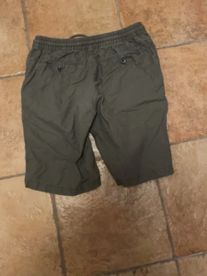 Mörkgröna shorts med snörning - Säljer ett par mörkgröna shorts med elastisk midja och snörning framtill. Shortsen har två bakfickor med knapp och är tillverkade i ett mjukt bomullsmaterial. Perfekta för varma dagar och enkel stil.