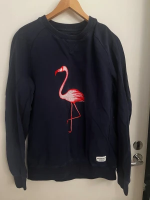 Mörkblå sweatshirt med flamingo - Mörkblå sweatshirt från Björn Borg med en stor rosa flamingo broderad på bröstet. Tröjan har rund halsringning, långa ärmar och ribbade muddar vid ärmslut och nederkant. Perfekt för dig som gillar sportig stil med en twist.