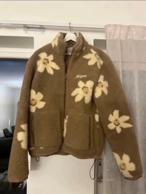 Brun teddyjacka med blommor från Axel Arigato - Supermysig brun teddyjacka med stora vita blommor och dragkedja framtill från märket Axel Arigato. Jackan har två stora fickor och hög krage. Perfekt för dig som vill sticka ut med en cool och trendig look. Materialet är fluffigt och ger en riktigt cozy känsla. 