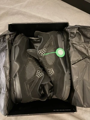 Svarta Air Jordan 4 Retro sneakers - Snygga helsvarta Air Jordan 4 Retro sneakers med ikoniska meshpaneler och detaljer i mocka. Skorna har en hög siluett, klassiska snörningar och Jumpman-logga på plösen. Perfekta för dig som vill ha en clean och stilren look.