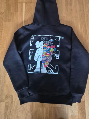 Svart  hoodie med tryck - Svart hoodie med stor färgglad grafisk print på ryggen och mindre tryck på bröstet. Klassisk huva med dragsko och magficka framtill. Materialet ser ut att vara mjuk bomullsblandning, perfekt för en streetwear-look. Strl S.... men är mer som M