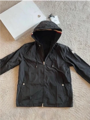 Svart vindjacka med huva - Snygg svart vindjacka med dragkedja framtill och två fickor med tryckknappar. Jackan har huva med dragsko och en diskret rand i rött och vitt längs kanten. Liten logotyp på ärmen. Tillverkad i lätt, vattenavvisande material. Boxen ingår inte!!