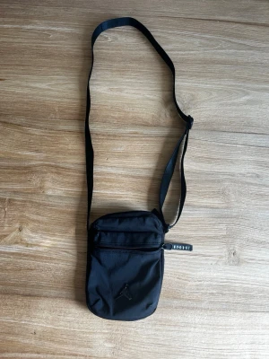 Svart axelväska med Jordan-logga - Snygg svart axelväska från Jordan med justerbar rem och dragkedja framtill. Väskan har en stilren design med den klassiska Jumpman-loggan på framsidan och är tillverkad i slitstarkt tygmaterial. Perfekt för att bära det viktigaste när du är på språng.