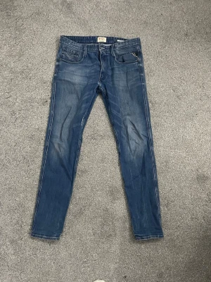 Replay Anbass  - Replay Anbass Slim Jeans – Feta slitningar – Strl 31    Snygga Replay Anbass i klassisk slim fit med riktigt feta, naturliga slitningar och whiskering – den där inkörda looken som är svår att hitta.  Storlek: 31 (midjemått) Modell: Anbass, slim fit Färg: Mellanblå denim med ljusare fades.   Bra begagnat skick. Två små hål har blivit lagade/sydda – det syns knappt och märks inte alls när man har dem på sig. Slitningarna är originella från replay när man köper vilket gör de här ännu feta re. 