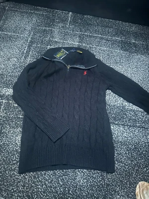 Svart kabelstickad tröja Polo Ralph Lauren - Kabelstickad svart tröja från Polo Ralph Lauren med halv dragkedja och röd broderad logga på bröstet. Tröjan har hög krage och ribbade muddar vid ärmslut och nederkant. Perfekt för lager på lager och riktigt snygg till jeans.
