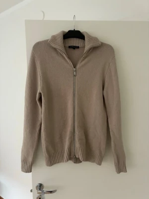 Beige ziptröja från Brand Island - Beige stickad ziptröja från Brand Island med hög ribbad krage och dragkedja framtill. Tröjan har långa ärmar och ribbade muddar vid ärmslut och nederkant. Perfekt för lager på lager och enkel att matcha med olika outfits.