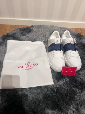 Valentino Garavani skor - Snygga vita sneakers från Valentino Garavani med en mörkblå rand och nitar på sidorna. Skorna har klassisk låg profil, vita snören och detaljer i gummi. Perfekta för dig som vill ha en exklusiv och trendig look.