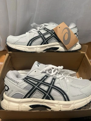 Asics skor - Snygga beige sneakers från Asics med svarta detaljer och logga på sidan. Skorna har en chunky sula, meshpaneler för bra ventilation och klassisk snörning. Perfekta för dig som gillar sportig och trendig stil.
