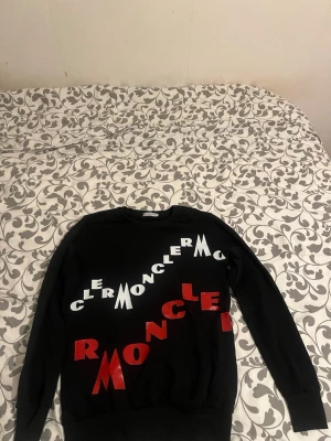 Svart sweatshirt med Moncler-tryck - Cool svart sweatshirt med stora vita och röda Moncler-bokstäver tryckta diagonalt över framsidan. Tröjan har rund hals och långa ärmar, perfekt för en streetwear-look.