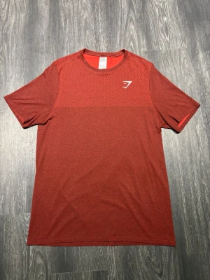 Gymshark Röd T-shirt 2016 - Äldre Gymshark Performance T-Shirt, ca 2016, i mörkröd/burgundy melerad design. Atletisk passform med ventilerande meshpanel över bröst och axlar för bra luftflöde under träning. Lätt och följsamt funktionsmaterial med klassisk vit Gymshark-logga på bröstet. Extremt fint skick, mycket välbevarad och nästintill som ny. Perfekt för gym och träning.