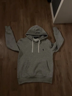 Ralph lauren hoodie grå - Snygg grå hoodie med vit snörning i huvan och klassisk känguruficka framtill. Liten broderad logga på bröstet. Perfekt för en avslappnad och stilren look.