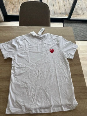Vit t-shirt med rött hjärta - Säljer en vit t-shirt med ett broderat rött hjärta med ögon på bröstet. Klassisk rund hals och korta ärmar. Tillverkad i mjuk bomull, perfekt för en clean och stilren look. Passar även M!