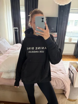 Svart hoodie Anine Bing Los Angeles - Säljer en svart hoodie från Anine Bing med vit text 'ANINE BING LOS ANGELES' på bröstet. Hoodien har en klassisk huva och stor magficka framtill. Perfekt oversized fit och mjukt material som känns skönt mot huden.