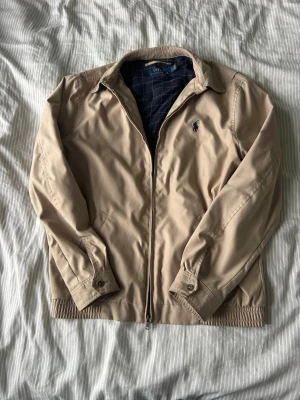 Beige vindjacka från Polo Ralph Lauren - Klassisk beige vindjacka från Polo Ralph Lauren med dragkedja framtill och broderad logga på bröstet. Jackan har krage, långa ärmar och ribbade muddar vid ärmslut och nederkant. Perfekt till vardags för en stilren look.Jackan är lite sliten i en av ärmarna men det syns knappast.Den passar personer runt 170 cm.