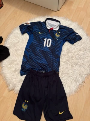 Frankrike fotbollströja Mbappé #10 - Säljer ett blått Frankrike fotbollsställ från Nike med Mbappé och nummer 10 på ryggen. Tröjan har vita detaljer, guldfärgade loggor och ett tupp-emblem på bröstet. Matchande blå shorts med Nike-logga och Frankrikes landslagsemblem. Perfekt för fotbollsträning eller match!