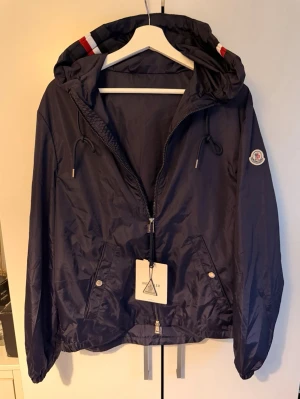 Moncler vindjacka Grimpeura - Snygg mörkblå vindjacka med dragkedja framtill och justerbar huva. Jackan har två fickor med tryckknappar och dragsko i nederkant. Tillverkad i lätt och glansigt material som passar perfekt för blåsiga dagar.