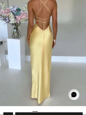 Ljusgul långklänning med slits - Snygg ljusgul långklänning i satin med smala axelband, öppen rygg med snörning och hög slits framtill. Klänningen har en figurnära passform och ett elegant, glansigt material som ger en lyxig känsla.